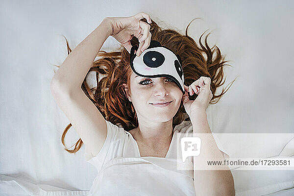 Smiling woman removing eye mask while lying on bed at home