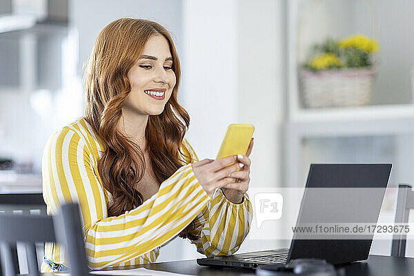 Lächelnde rothaarige Geschäftsfrau  die ein Smartphone benutzt  während sie zu Hause am Laptop sitzt