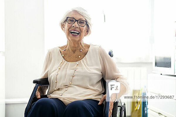 Fröhliche behinderte Seniorin mit Brille  die zu Hause im Rollstuhl sitzt