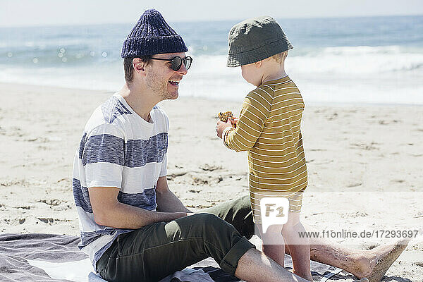 Lächelnder Vater sieht seinen Sohn beim Essen am Strand an