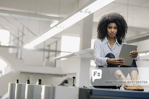 Businesswoman with digital tablet sitting on machinery at industry