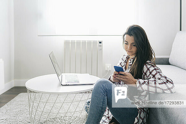 Junge Frau mit Laptop  die ein Mobiltelefon benutzt  während sie im Wohnzimmer sitzt