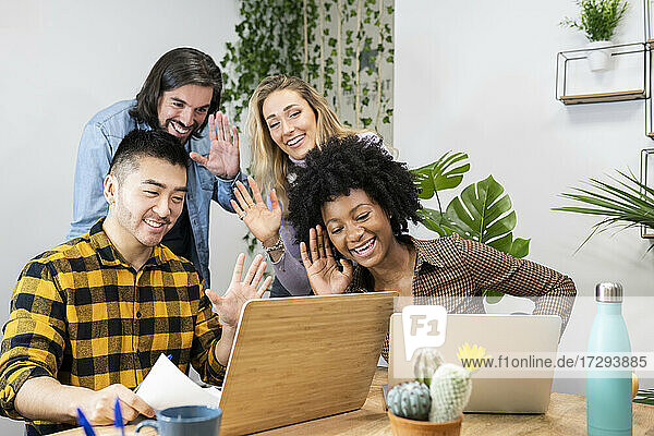 Lächelnde männliche und weibliche Fachkräfte winken bei einer Videokonferenz über einen Laptop im Büro