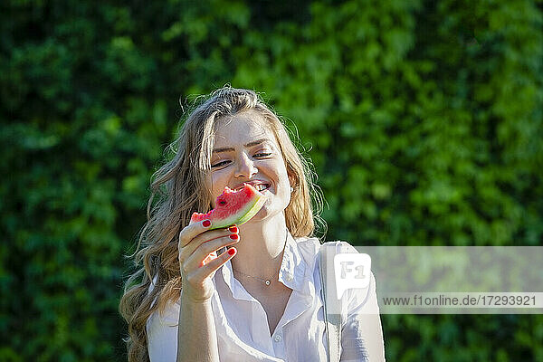 Lächelnde junge Frau mit Wassermelonenscheibe im Garten an einem sonnigen Tag