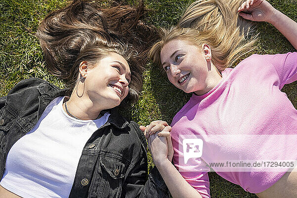 Lächelnde Freundinnen  die sich an den Händen halten und sich gegenseitig ansehen  während sie im Park auf dem Rasen liegen