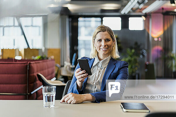 Lächelnde weibliche Fachkraft sitzt mit Handy am Schreibtisch im Büro
