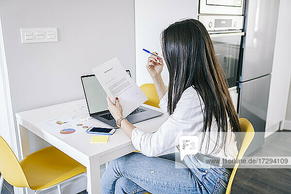 Young female freelance worker reading document while sitting at home office