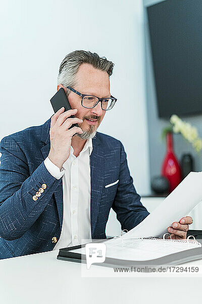 Männlicher Unternehmer prüft ein Dokument  während er im Büro mit dem Handy telefoniert