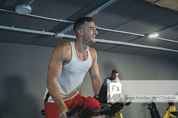 Male athlete smiling while cycling with friend in background at gym