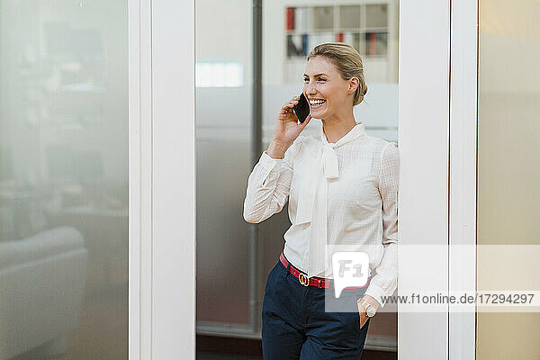 Fröhliche weibliche Fachkraft  die mit ihrem Smartphone spricht  während sie an der Tür im Büro lehnt