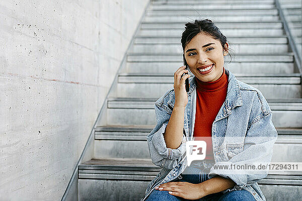 Lächelnde Frau in Jeansjacke  die im Treppenhaus mit ihrem Smartphone spricht