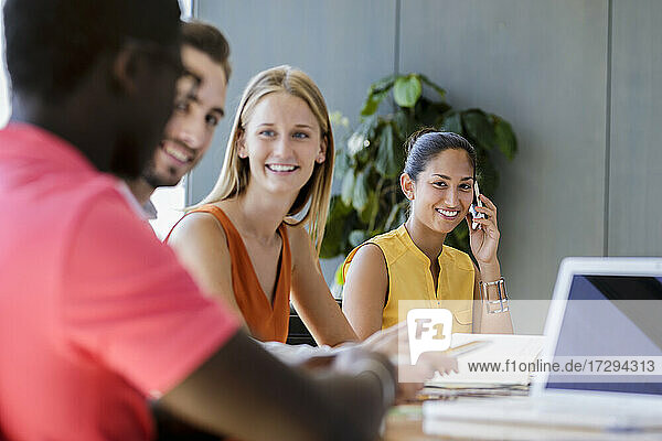 Lächelnde Geschäftsfrau  die auf einem Smartphone spricht  während männliche und weibliche Unternehmer in einer Besprechung im Büro diskutieren