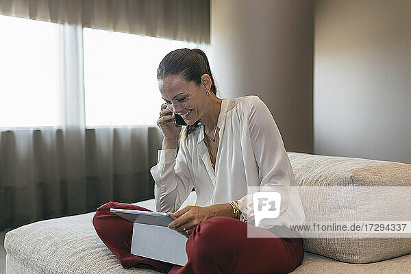 Glückliche reife Geschäftsfrau,  die mit einem digitalen Tablet auf dem Sofa sitzt und telefoniert