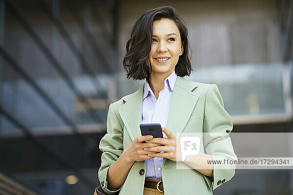 Smiling businesswoman looking away while holding smart phone outside office building