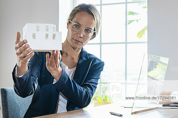 Architektin mit Hausmodell sitzt vor einem futuristischen Bildschirm im Büro