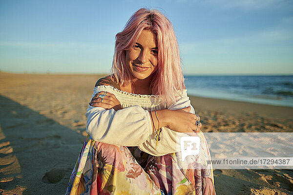 Lächelnde junge Frau mit verschränkten Armen  die bei Sonnenuntergang am Strand sitzt