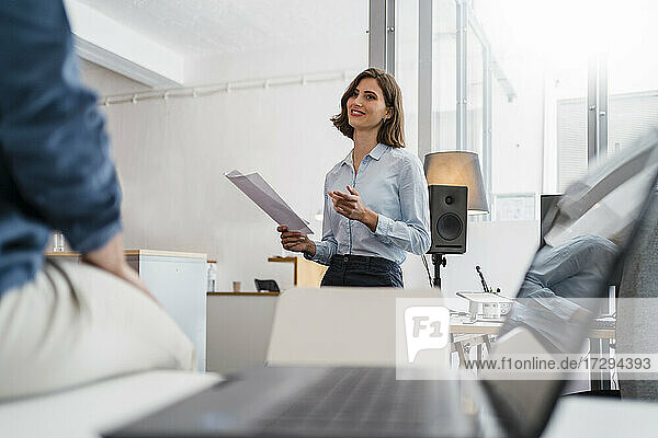 Junge Geschäftsfrau hält Dokumente in der Hand  während sie mit einem Kollegen im Büro diskutiert