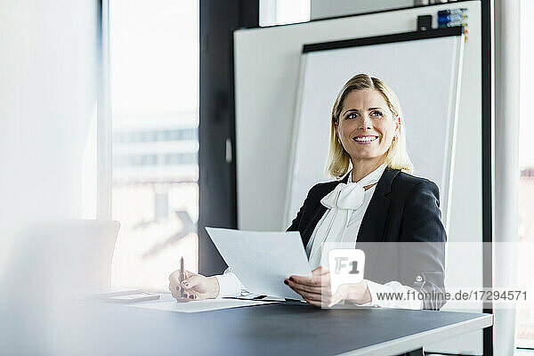 Lächelnde Unternehmerin mit Dokumenten am Schreibtisch sitzend und wegschauend