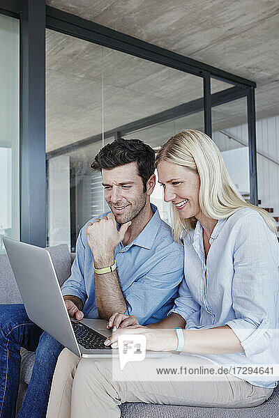 Lächelndes Paar im mittleren Erwachsenenalter  das einen Laptop benutzt und zu Hause auf dem Sofa sitzt