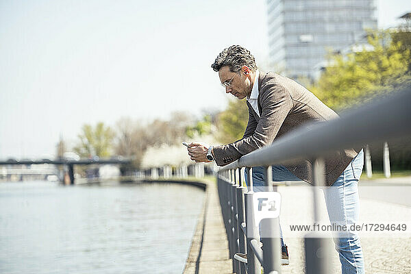 Älterer Geschäftsmann  der ein Mobiltelefon benutzt  während er sich auf ein Geländer am Fluss stützt