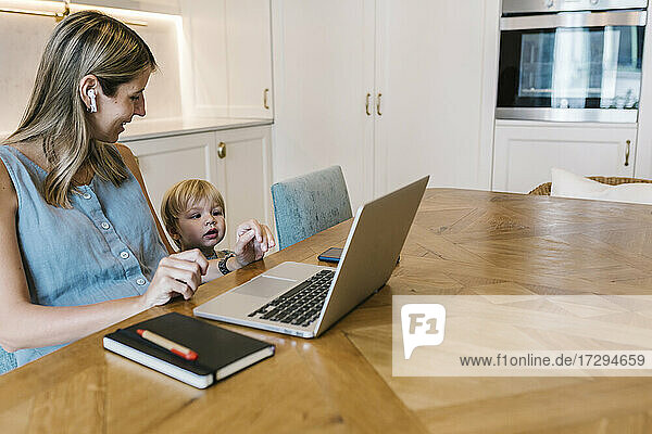 Lächelnde Freiberuflerin mit Blick auf den neugierigen Sohn vor dem Laptop am Esstisch in der Küche
