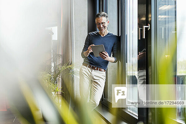 Fröhlicher Geschäftsmann  der ein digitales Tablet benutzt und sich an ein Glasfenster im Büro lehnt