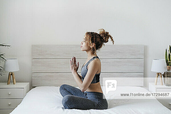 Junge Frau mit verschränkten Händen meditiert auf dem Bett zu Hause