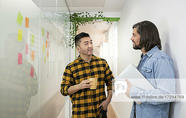 Männlicher Berufstätiger  der einen Kollegen ansieht  der mit einem Laptop steht und diskutiert  während er im Bürokorridor steht