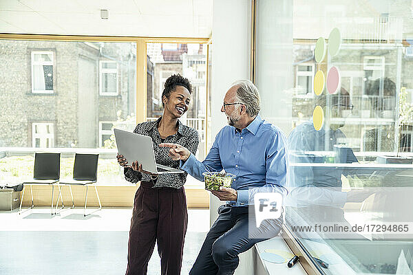 Lächelnde männliche und weibliche Kollegen  die sich gegenseitig ansehen  während sie über einen Laptop im Büro diskutieren