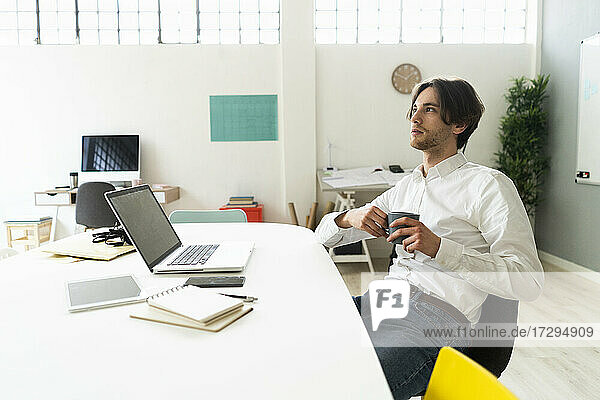 Nachdenklicher Geschäftsmann mit Kaffeetasse am Schreibtisch sitzend im Büro