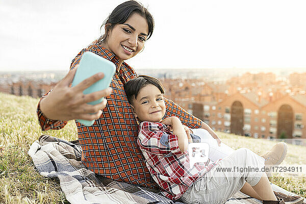 Lächelnde Frau  die ein Selfie mit einem Jungen macht  während sie auf einem Hügel sitzt