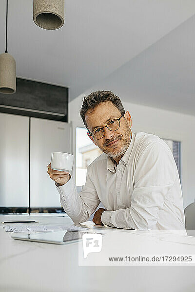 Geschäftsmann mit Brille  der eine Kaffeetasse an der Kücheninsel im Heimbüro hält