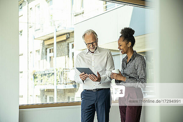 Smiling male and female colleagues looking at digital tablet while standing in office