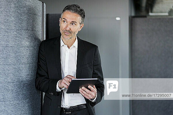 Mature businessman with digital tablet standing by wall while looking away