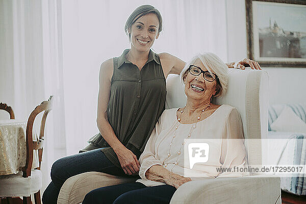 Lächelnde ältere Frau sitzt mit ihrer Enkelin auf einem Sessel im Wohnzimmer zu Hause
