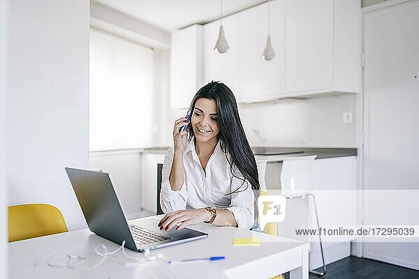 Female freelance worker with laptop talking on mobile phone at home office