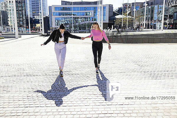 Weibliche Freunde  die sich an den Händen halten und ihren Schatten betrachten  während sie an einem sonnigen Tag auf einem Fußweg spazieren gehen