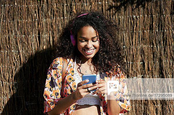 Lächelnde Frau mit Smartphone vor einer Strohwand an einem sonnigen Tag