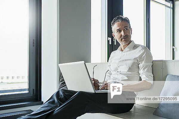 Männlicher Berufstätiger sitzt mit Laptop auf Möbeln im Büro und schaut weg