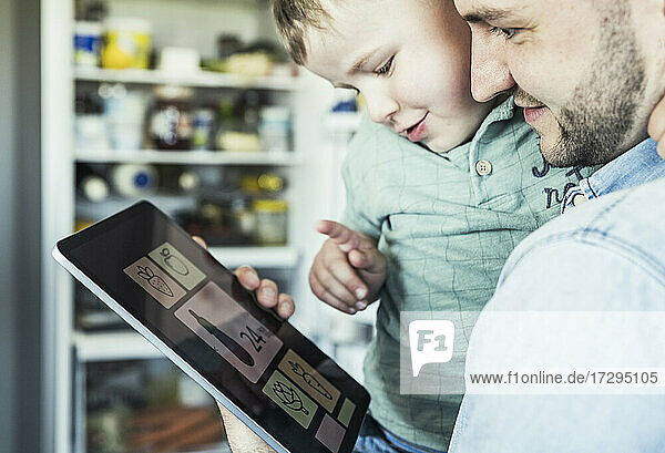 Lächelnder erwachsener Mann mit Sohn  der in der Küche auf ein digitales Tablet zeigt
