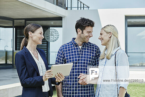 Immobilienmaklerin mit digitalem Tablet  die ein lächelndes Paar ansieht
