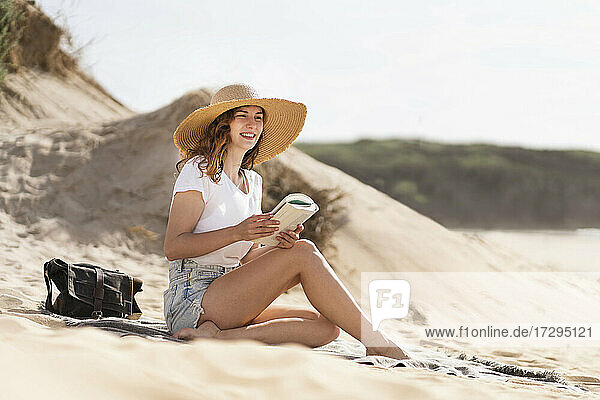 Lächelnde junge Frau mit Hut sitzt mit Buch an einem sonnigen Tag am Strand während der Ferien