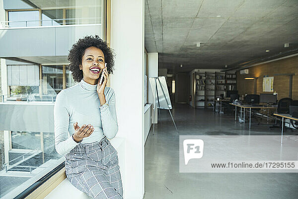 Weibliche Fachkraft gestikuliert  während sie mit ihrem Smartphone in der Nähe eines Glasfensters im Büro spricht