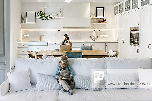 Kleiner Junge telefoniert auf dem Sofa  während die Mutter im Hintergrund in der Küche arbeitet