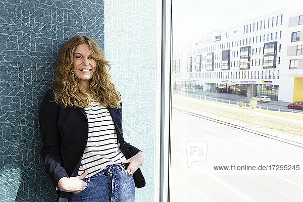 Weibliche Unternehmerin lächelt  während sie sich im Büro an die Wand am Fenster lehnt