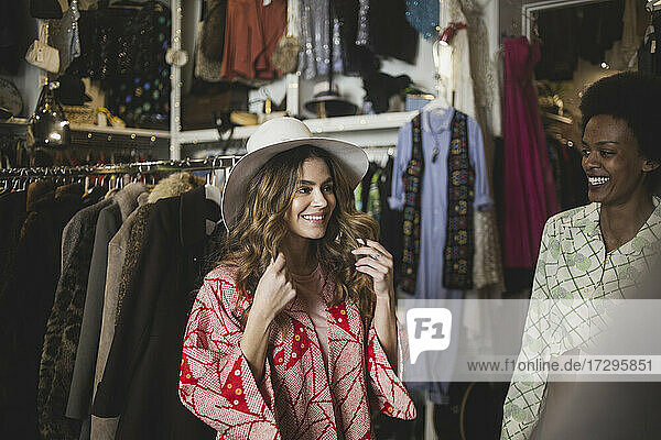 Smiling woman trying on hat while standing by female owner in clothing store