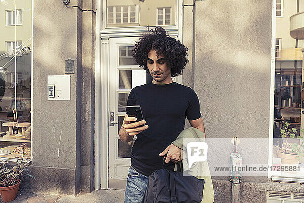 Mid adult man with curly hair using smart phone while standing outside cafe