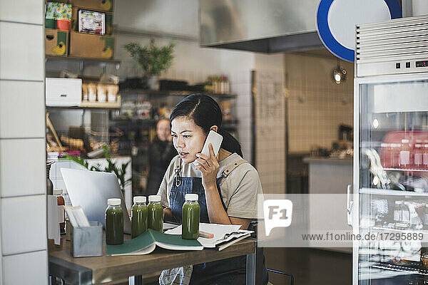 Asiatische weibliche Unternehmerin spricht über Smartphone während der Arbeit am Laptop im Laden
