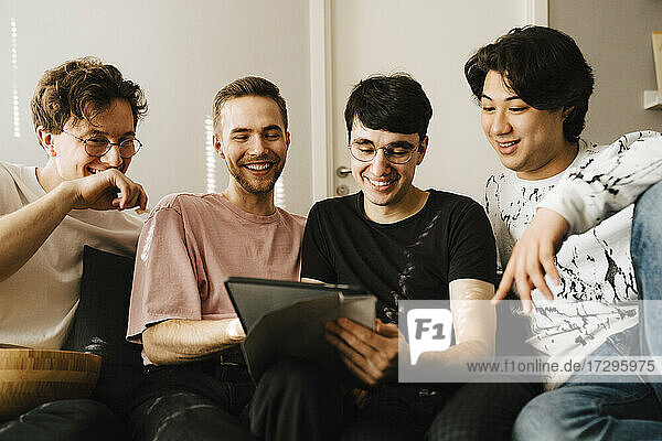 Lächelnder Mann  der ein digitales Tablet benutzt  während er sich mit männlichen Freunden im Wohnzimmer austauscht