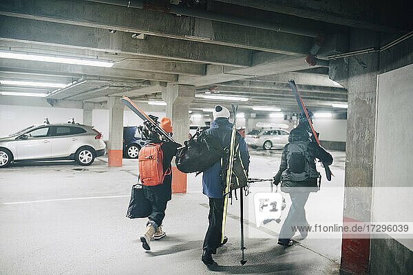 Full length rear view of male and female friends carrying skis while walking at parking garage